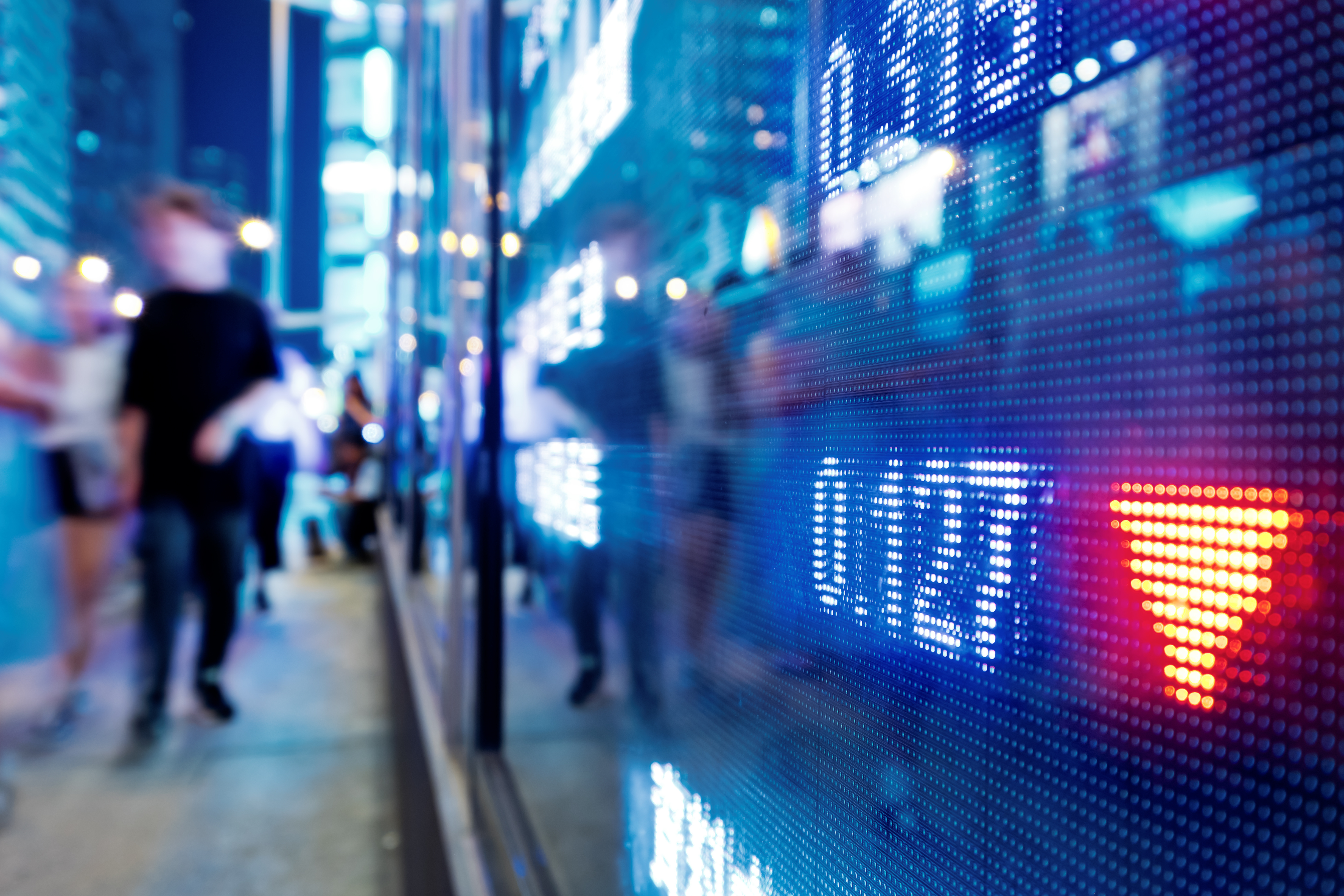 Close-up of a vibrant blue and red digital stock price display, with blurred pedestrians walking by in the background.