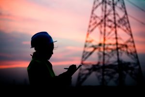 Silhouette of an engineer wearing a safety helmet and reflective vest, writing on a clipboard near a power transmission tower at sunset.