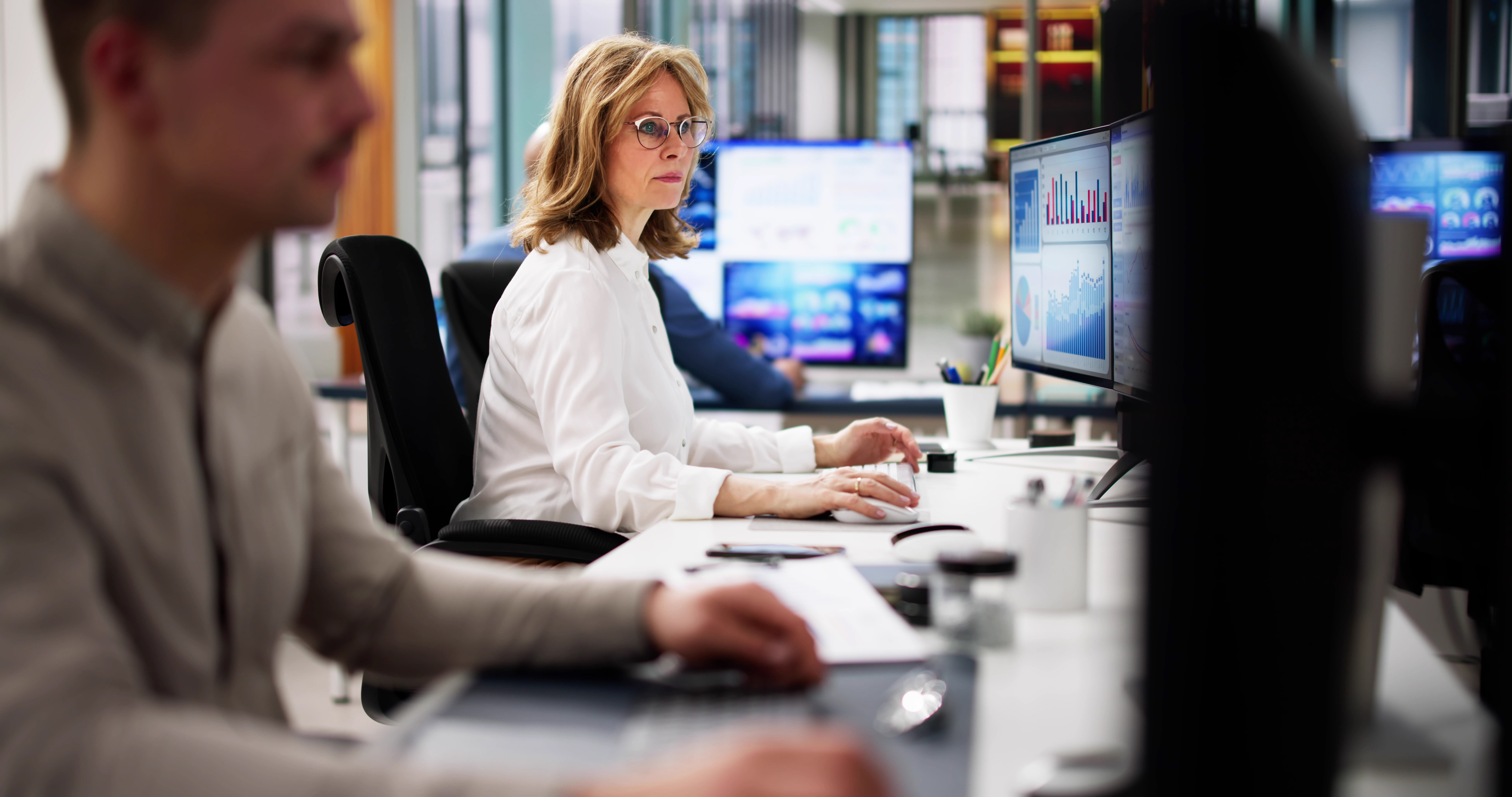 Two professionals seated at desks in an office; a woman in the middle ground intently uses a computer while a man is visible in the foreground.
