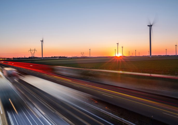 Cars speed along a highway at sunset with wind turbines spinning across open fields in the background.